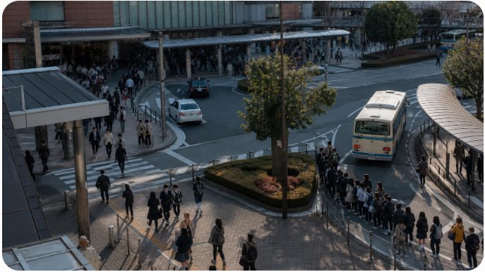 A busy urban plaza with pedestrians crossing a street while a bus travels along a curved road near a shopping area.